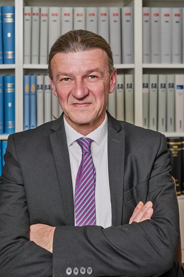 Foto von Andreas Vogtmann vor der Bücherwand sitzend an einem Tisch mit Blick in die Kamera