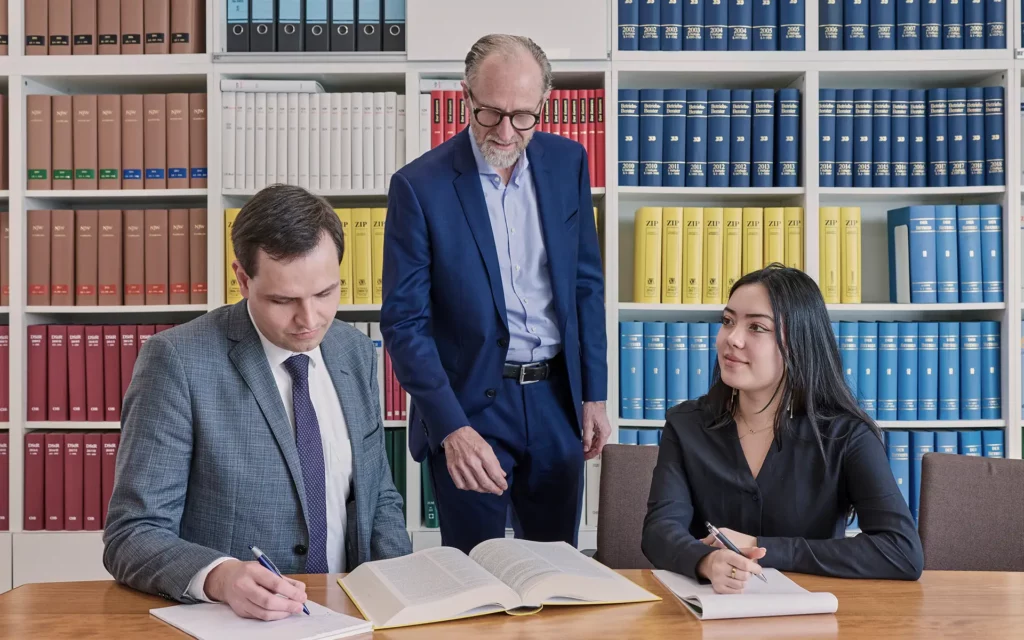 Zwei Personen sitzen am Tisch und notieren Inhalte aus einem aufgeschlagenen Fachbuch, eine weitere Person steht daneben, Hintergrund mit juristischer Fachliteratur