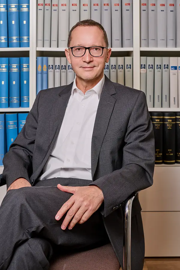 Foto von Jochen Jahn vor der Bücherwand sitzend an einem Tisch mit Blick in die Kamera