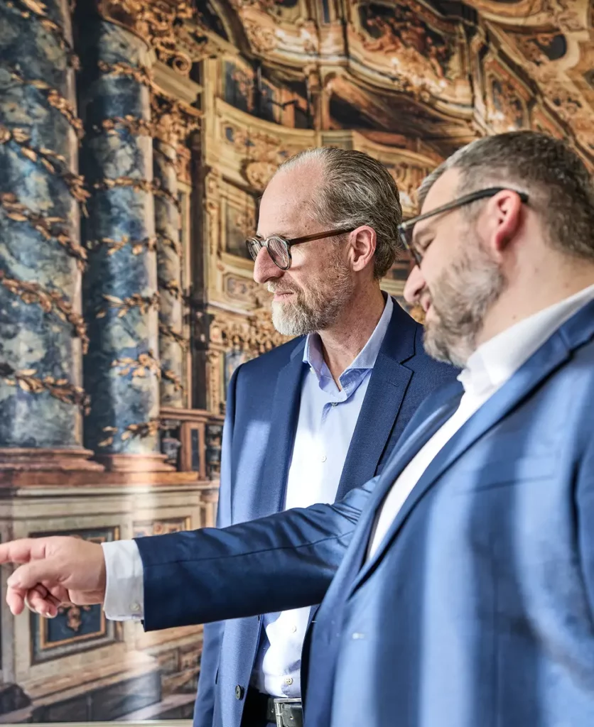 Dr. Alfred Krammer und Stefan Behrendt im Anzug betrachten gemeinsam etwas außerhalb des Bildes, Stefan Behrendt zeigt mit der Hand, im Hintergrund ein kunstvolls Bild, der Oper Bayreuth