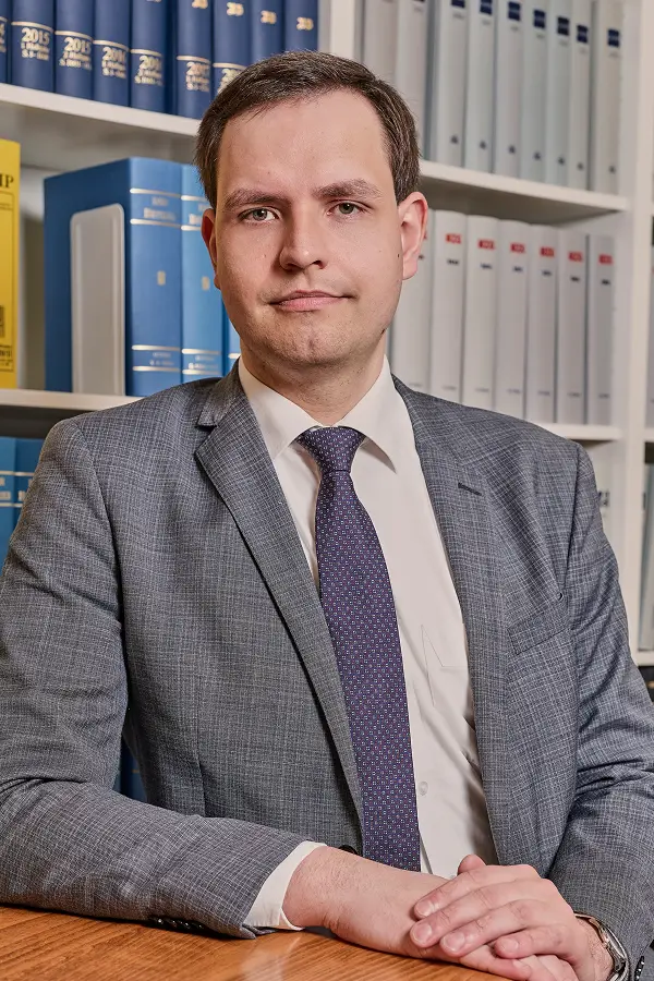 Foto von Thomas Loos vor der Bücherwand sitzend an einem Tisch mit Blick in die Kamera