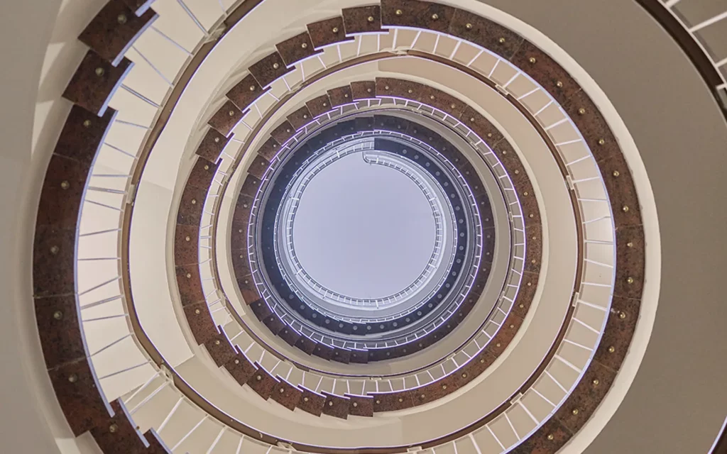 Das Treppenhaus von der Kanzlei. Es ist eine Treppe, die im Kreis verläuft. Das Foto wurde vom Erdgeschoss aufgenommen und blickt mittig bis an die oberste Decke im Gebäude